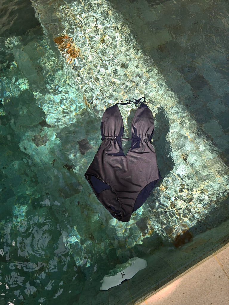Sun Kits Billie One Piece in Black floating in a pool with crysatl clear water and turquoise green tiles. The sun is shining on the swimsuit. 
